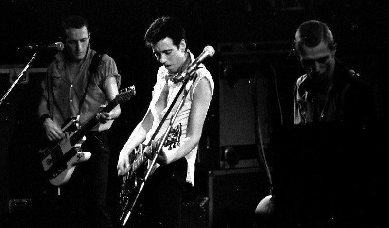 The Clash tocando en vivo en 1980, con Joe Strummer al micrófono y guitarra, Paul Simonon en el bajo y Mick Jones en la guitarra, durante un show intenso y cargado de energía punk