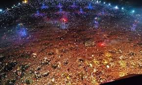 Vista aérea nocturna de una multitud masiva en un evento al aire libre, iluminada por luces de colores y rodeada de reflectores.