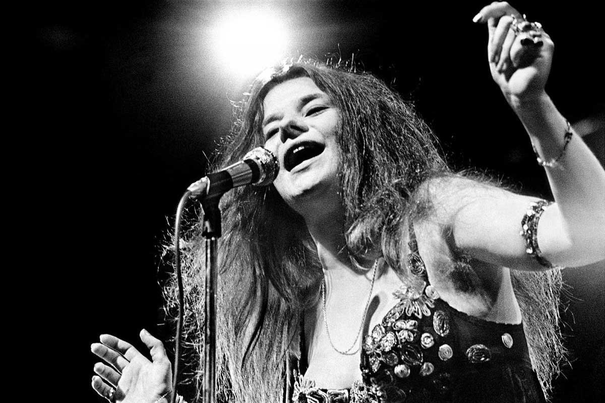 Janis Joplin sonriendo en una foto en blanco y negro, con su estilo hippie característico, pulseras y cabello despeinado, ícono del rock de los años 60